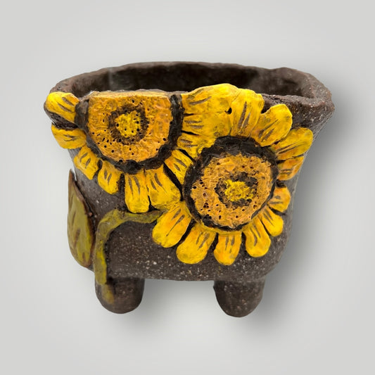 This small, four-legged planter exudes rustic charm with its textured brown surface. The standout feature is the three-dimensional sunflower motifs with vibrant yellow petals and brown centers, which add a delightful pop of color and artistic flair. These sunflowers create a naturalistic and eye-catching design, making this planter a perfect addition for anyone looking to bring a touch of floral elegance to their home or garden decor.