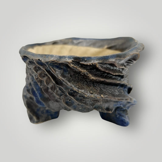 This uniquely crafted pot features an irregular, wavy rim and a textured exterior that resembles natural stone or geological formations. The color palette ranges from deep blues to subtle grays, mimicking the hues of minerals or the ocean depths. 