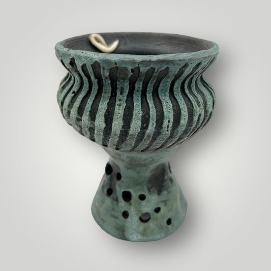 This handcrafted ceramic planter features a unique goblet shape. The wide, rounded bowl sits atop a narrower pedestal base. The glaze work is exquisite, combining glossy and matte finishes in shades of teal and dark gray. The exterior of the bowl showcases an intricate pattern of vertical ridges and carved lines, adding texture and visual interest.

The circular cut-outs on the pedestal base not only enhance the aesthetic but also serve a practical purpose—they allow for better air circulation around the pl