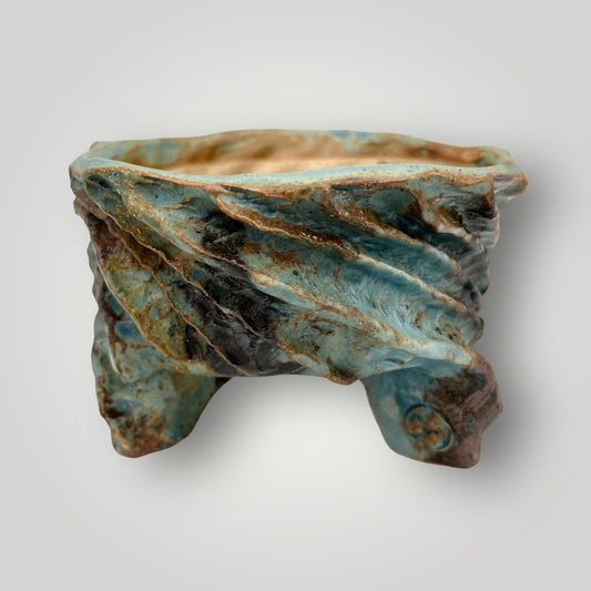 This uniquely crafted ceramic pot is a stunning piece that combines functionality with artistic flair. It features a wide, open top that tapers down to a base supported by three rounded feet. The glaze showcases a blend of earthy tones, predominantly in shades of turquoise and brown with hints of cream.