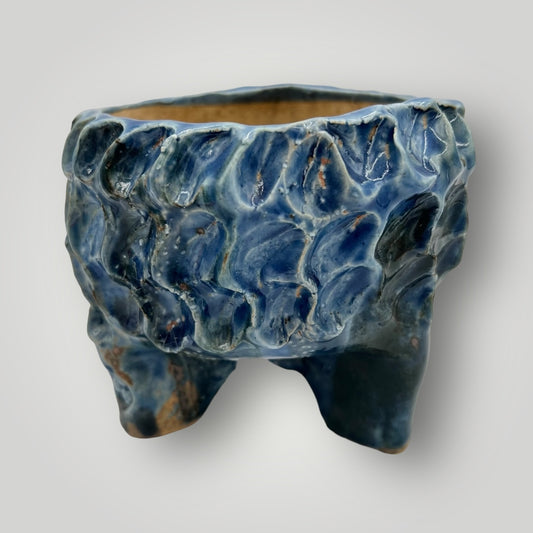 The pot in your image is a beautiful ceramic piece with a unique, irregular shape. Its textured surface resembles flowing or rippling water, featuring various shades of blue with hints of white, giving it an appearance reminiscent of ocean waves.